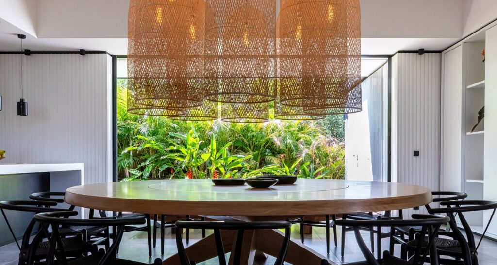Round wood dining table under woven pendant lights beside lush garden, Villa Valentina, St. Barts.
